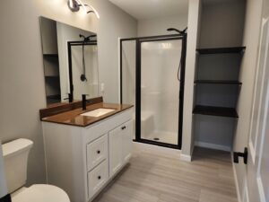 Master bathroom addition in Elizabethtown, PA with wood flooring, a white vanity with brown countertop, black framed shower, and black shelving.
